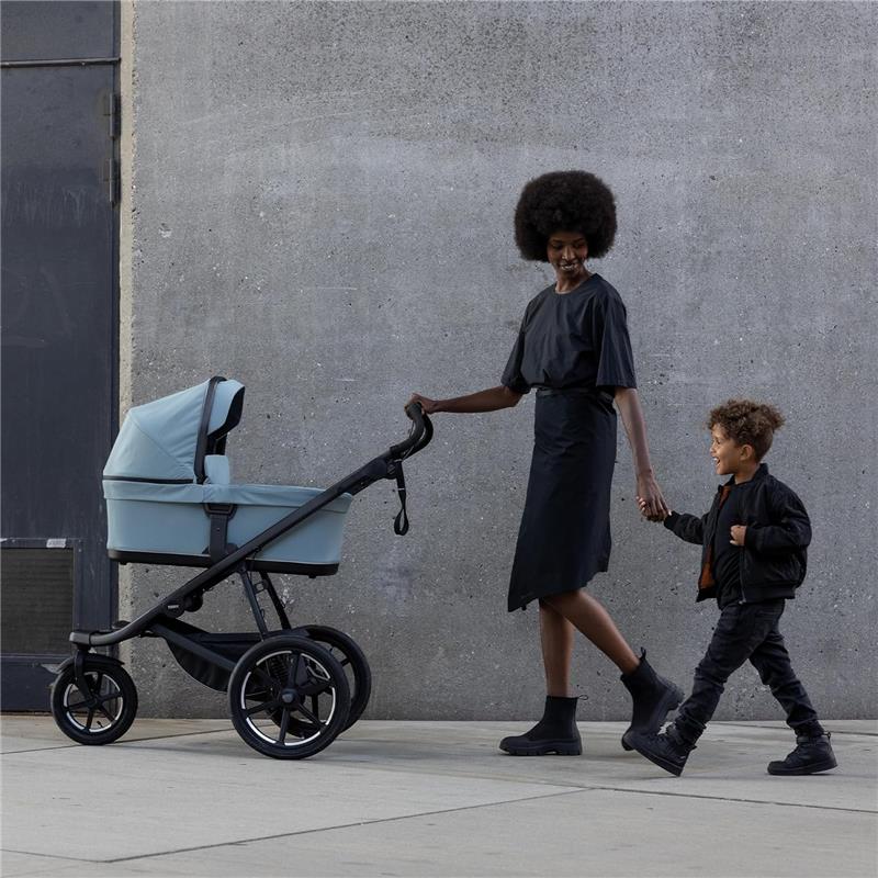 A woman in a black dress pushes a blue Thule Bassinet Mid stroller while holding hands with a smiling child.