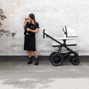A woman in a black dress stands beside a Thule bassinet in soft beige, holding a baby. The bassinet is on a stroller with black wheels.