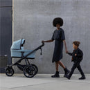 A woman in a black outfit pushes a soft beige Thule bassinet stroller, holding hands with a smiling child.