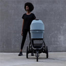Thule Bassinet in soft beige, positioned on a stroller, with a standing person wearing a black outfit in an outdoor setting.