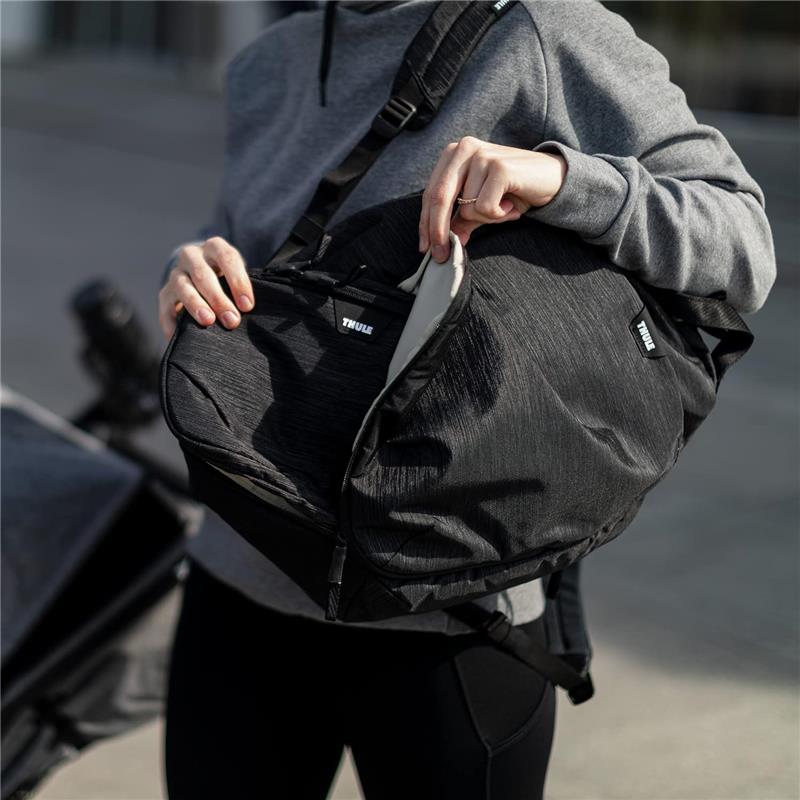 A person wearing a gray sweatshirt holds a Thule black changing backpack, revealing its inner compartment.