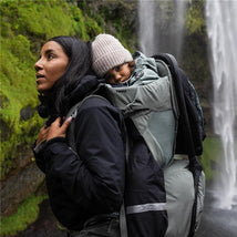 A woman carries a child in a Thule Sapling Child Carrier Backpack while standing near a waterfall and greenery.