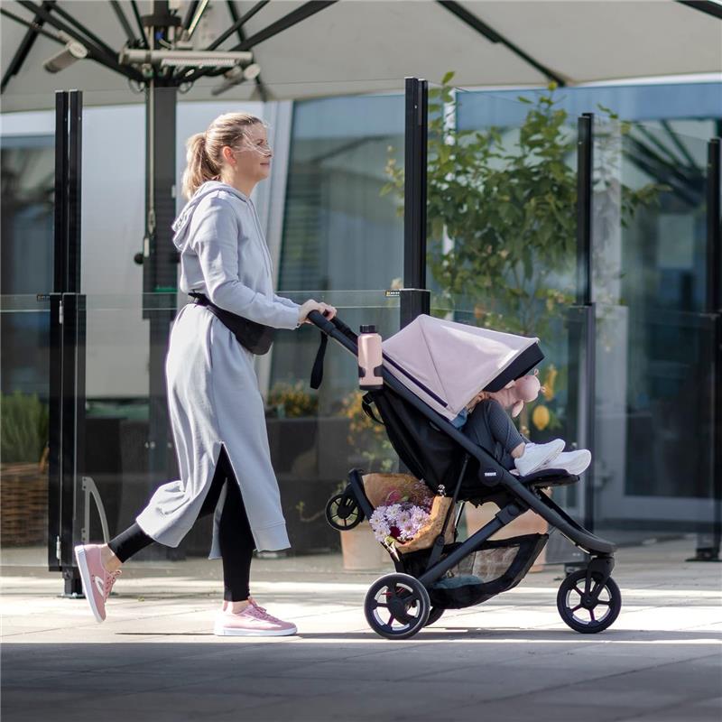 Woman walking with a Thule stroller featuring a seated child, a water bottle holder, and a floral arrangement in the storage basket.