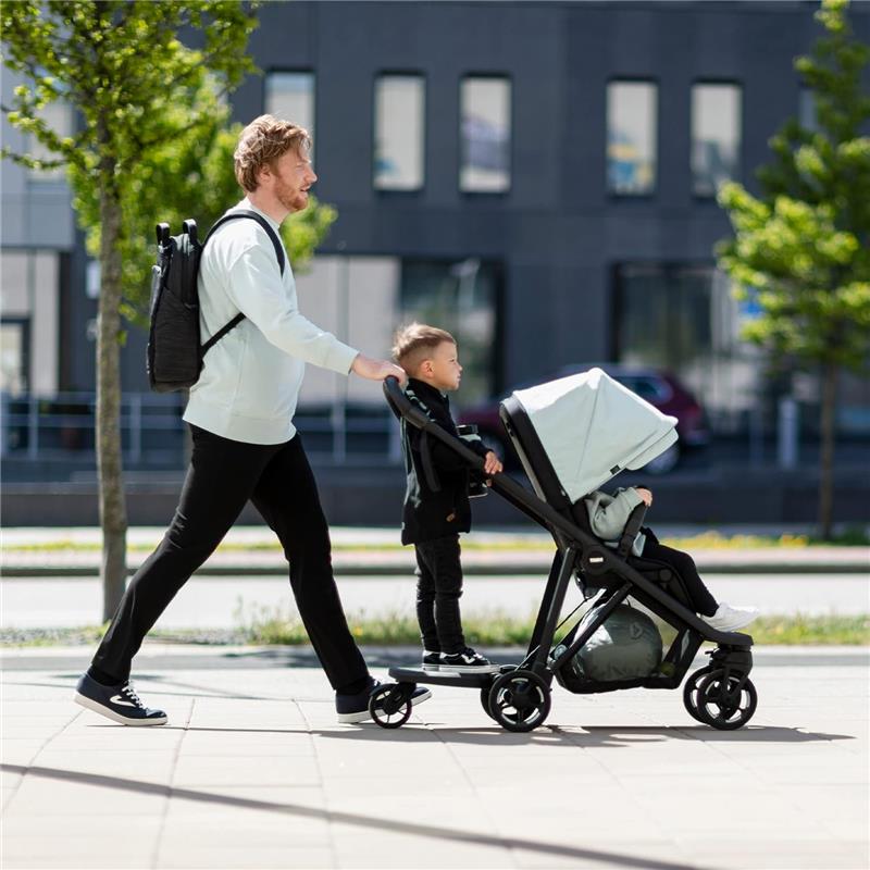A man walks outside, pushing a Thule stroller with a child seated inside, while another child stands on a rider board attached to the stroller.