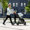 A man walks outside, pushing a Thule stroller with a child seated inside, while another child stands on a rider board attached to the stroller.