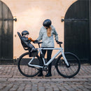 A woman and a child in helmets stand by a light gray bike with a Thule Yepp 2 Maxi rack-mounted seat.