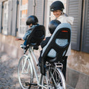 A woman smiles while standing next to a bicycle with two child seats mounted on the back, both equipped with helmets.
