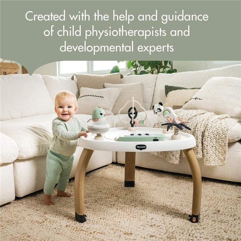 A baby stands beside the Tiny Love 5-in-1 Here I Grow Activity Center featuring black and white toys, with a cozy sofa in the background.