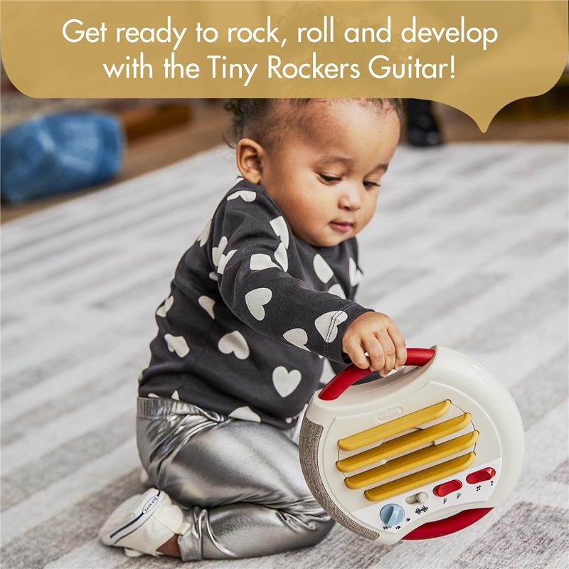 A toddler in a heart-patterned shirt plays with the Tiny Love Tiny Rockers Guitar, a colorful musical toy featuring buttons and a handle.
