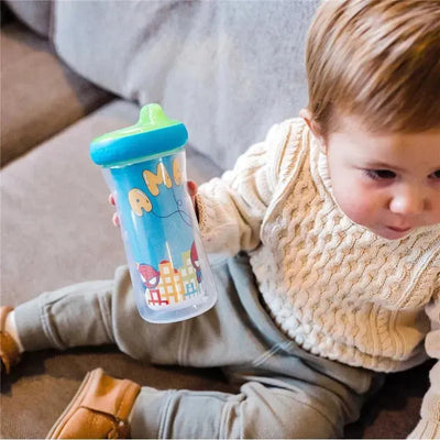 Toddler holding a 9oz Tomy Marvel Insulated Sippy Cup with colorful designs and a blue lid while sitting on a couch.