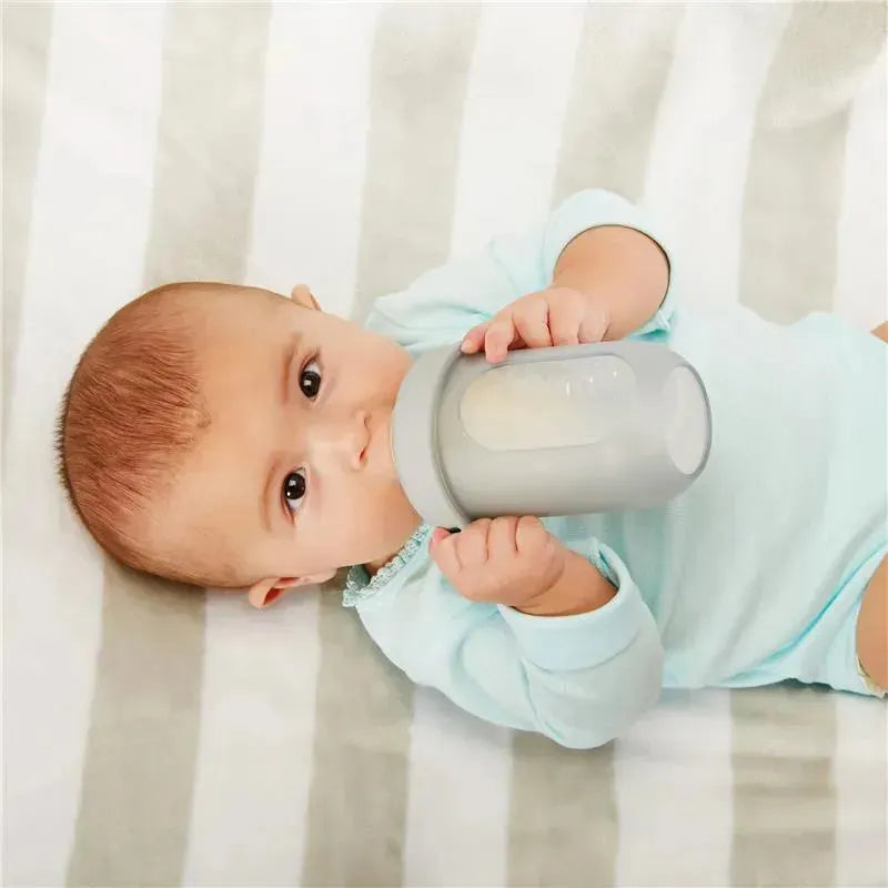 Baby holding a light gray Tomy Boon silicone bottle, lying on a striped blanket, wearing a blue onesie.