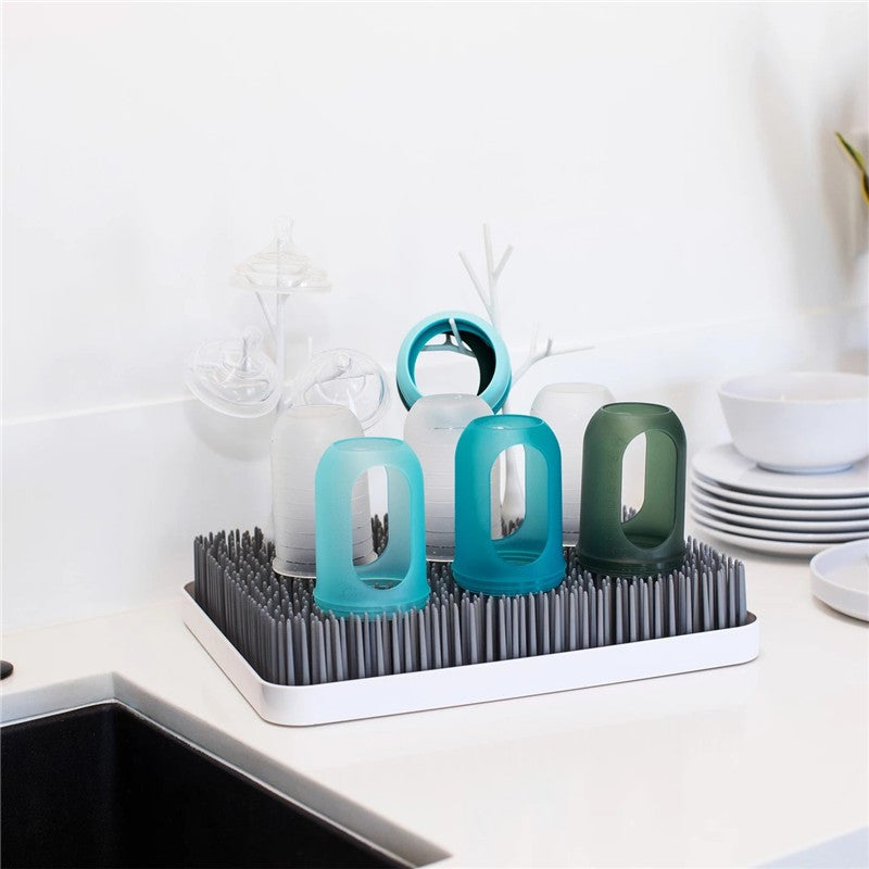 Boon LAWN drying rack displaying colorful cups and TWIG accessories on a countertop beside white dishware.