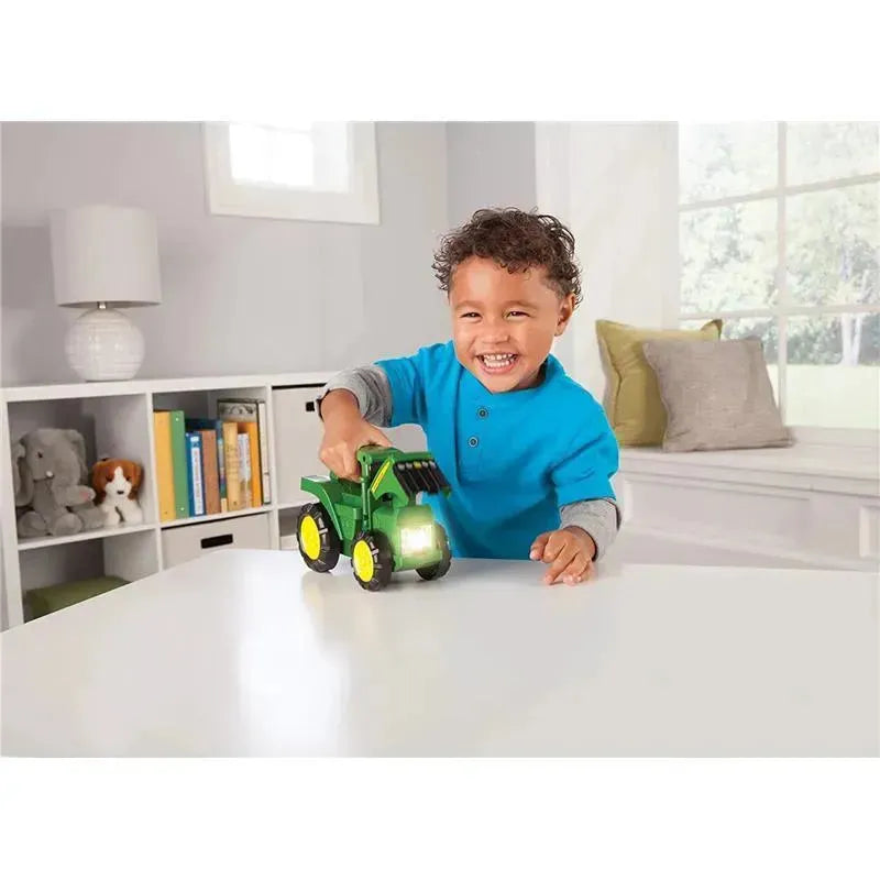 Smiling child in a blue shirt playing with a green John Deere Roll N Go flashlight toy on a white table.