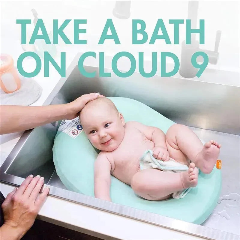 Baby enjoying a bath on a light green Puff Inflatable Bather in a sink, with adult hand supporting its head.