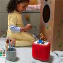 A young girl in yellow pajamas paints a cardboard box while a Tonies Bullseye audio toy figurine sits atop a red speaker.