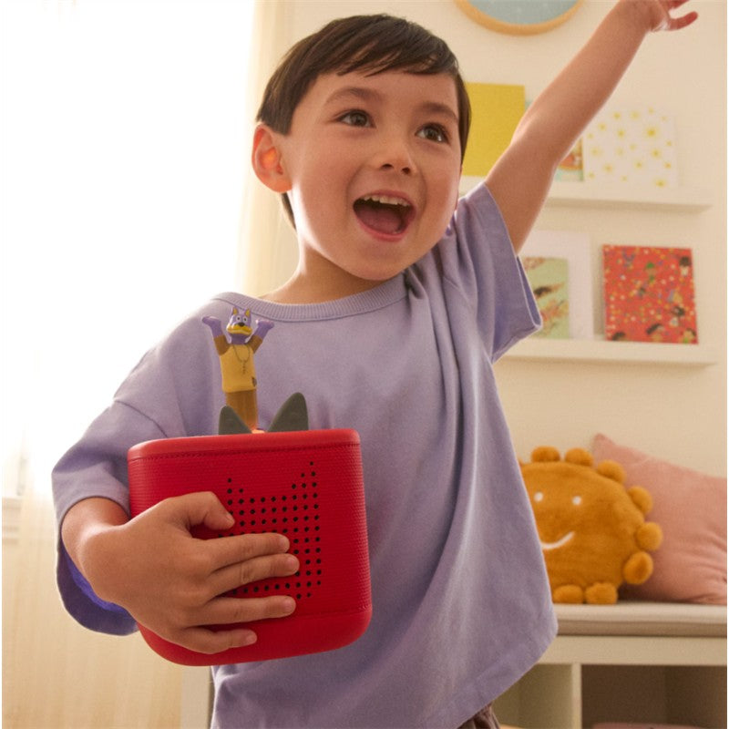 A joyful child holds a red Tonies Doggyland audio toy figurine with a character on top, smiling and raising their arm energetically.