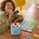 Child with curly hair holds a green Toniecard, smiling at a blue Toniebox on a wooden table, surrounded by colorful game cards and a map.