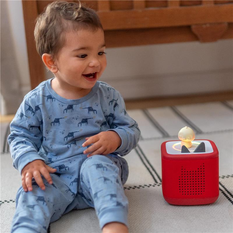 A smiling toddler in blue pajamas sits cross-legged next to a red Tonies Sleepy Duck audio toy, which features a small duck figurine.