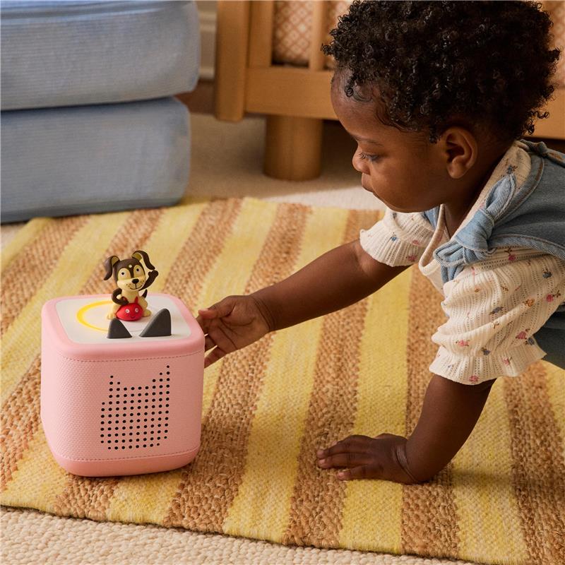 A child interacts with a pink Toniebox audio player featuring a playful puppy figure on top, set on a striped rug.