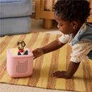 A child interacts with a pink Toniebox audio player featuring a playful puppy figure on top, set on a striped rug.