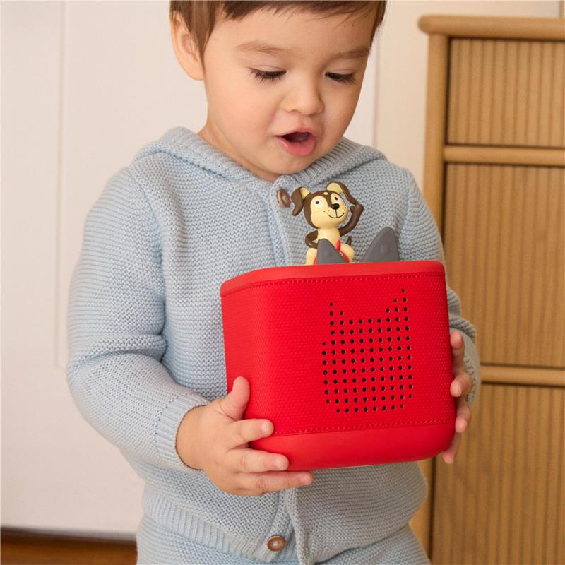 A toddler in a light blue sweater holds a red Toniebox audio player, featuring a Playtime Puppy character on top.