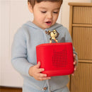 A toddler in a light blue sweater holds a red Toniebox audio player, featuring a Playtime Puppy character on top.