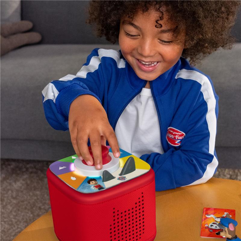 A child with curly hair smiles while pressing a button on the red Tonies Tonieplay Game Controller, featuring colorful icons on top.