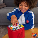 A child with curly hair smiles while pressing a button on the red Tonies Tonieplay Game Controller, featuring colorful icons on top.