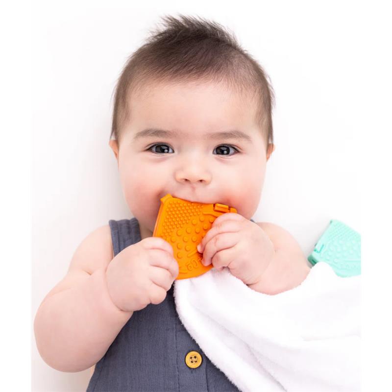 Baby holding an orange rubber teether clip, chewing on it while clasping a white towel.