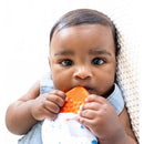 Baby holding an orange textured teether clip, looking directly at the camera, with a soft blanket in the background.