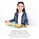 Girl with curly hair and glasses smiles while holding a plate of apple slices and sandwich on a green Ubbi Platemate.