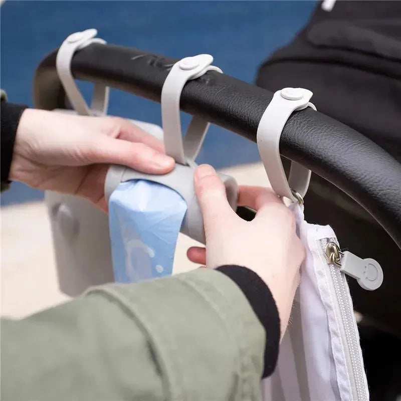 Hands attaching the Ubbi Retractable On the Go Bag Dispenser to a stroller handle, showcasing its design and functionality.