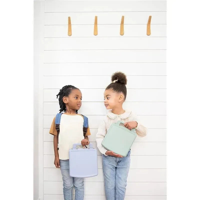 Two kids smile at each other while holding colorful bento lunch boxes; one is sage green and the other light blue.