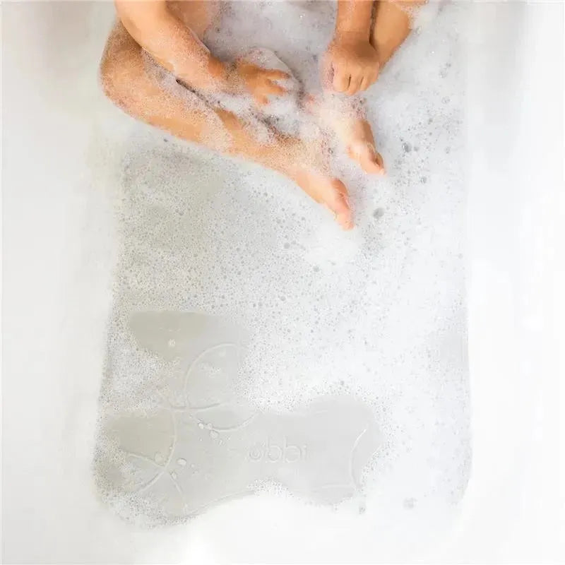 Cushioned, gray Ubbi non-slip bath mat in a bubbly bathtub with a baby’s feet visible.