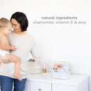 A woman holds a baby while reaching for Ubbi fragrance-free baby wipes from a white container on a white surface.