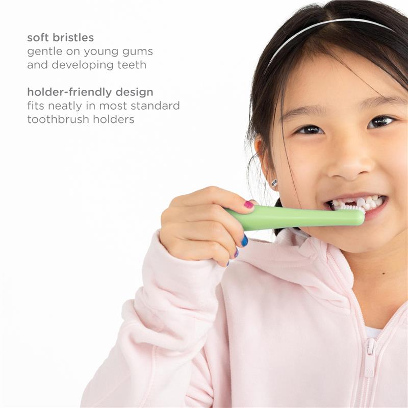 A young girl brushes her teeth with a green Ubbi training toothbrush featuring soft bristles, smiling while wearing a pink hoodie.