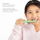 A young girl brushes her teeth with a green Ubbi training toothbrush featuring soft bristles, smiling while wearing a pink hoodie.