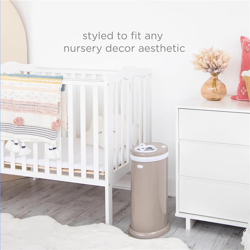 Stainless steel Ubbi diaper pail in taupe, designed for odor locking, next to a white crib and dresser in a styled nursery.