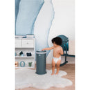 A toddler in a diaper interacts with a gray Ubbi Steel Diaper Pail, designed to lock odors without special bags, in a stylish nursery.