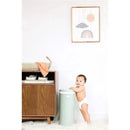 Sage green Ubbi Steel Odor Locking Diaper Pail next to a smiling baby in a diaper, with a wooden cabinet and decorative wall art.