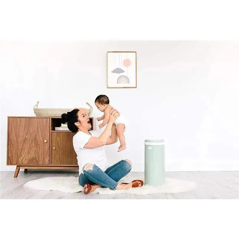 A woman in a white shirt joyfully holds a baby while sitting on a rug, with an Ubbi steel diaper pail in sage green nearby.