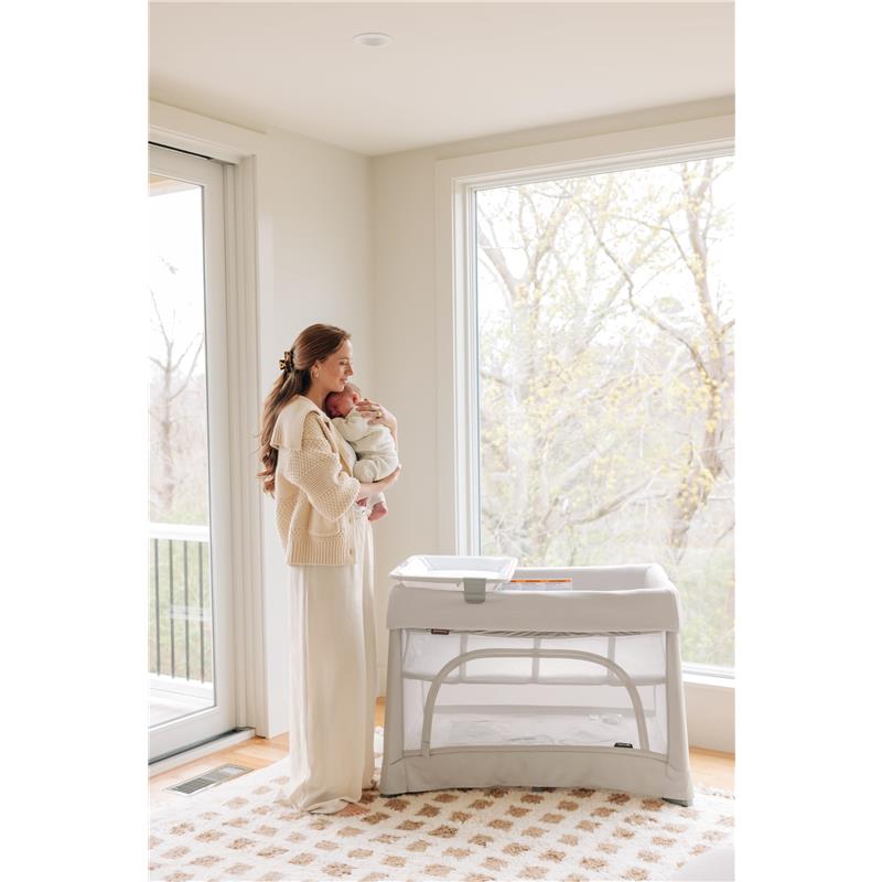 Mother holding a baby in a cozy room next to a UPPAbaby Remi V2 Playard, featuring a waterproof mattress cover.