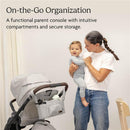 Carrying the UPPAbaby Carry-All Parent Organizer in charcoal, a woman holds her smiling child beside a stroller, showcasing its compartments.