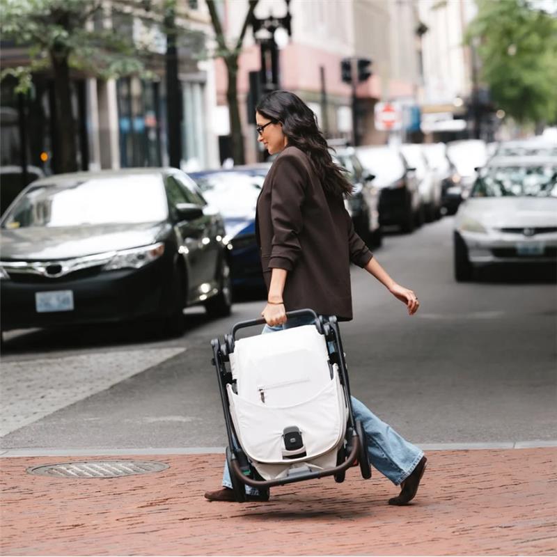 A woman wearing a brown blazer and jeans walks on a city street, carrying a folded Uppababy Kona stroller in black and white.