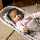 A baby in a pink sweater sits in an Uppababy Mira 2-in-1 bouncer, looking curiously at the viewer.
