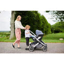 Uppababy Piggyback for Cruz V2 is attached to a stroller, with a woman pushing the stroller and a child standing on the board.