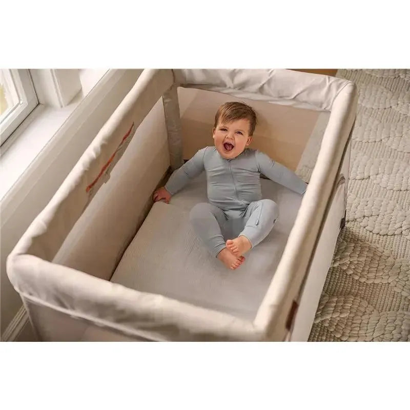 Infant wearing a light blue bodysuit, sitting in a cozy crib with a white mattress cover, joyfully smiling.