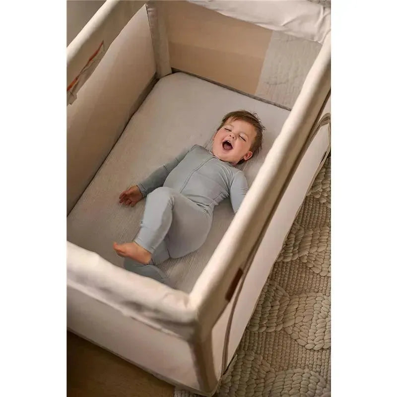 Child in light gray pajamas happily lying on the Uppababy Remi organic cotton mattress cover in a crib.