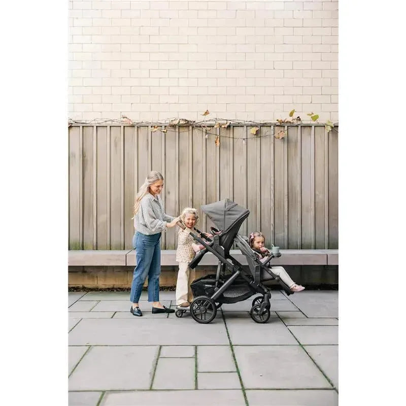 A woman pushes an Uppababy RumbleSeat V2+ stroller with two children inside, smiling, in a modern outdoor setting.