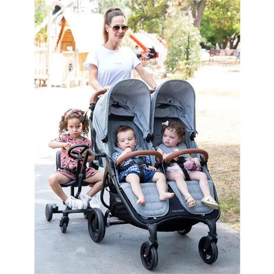 A woman pushes a Valco Baby Co-Rider Ride-Along Seat stroller with two children seated, while another child rides a small push-along toy.